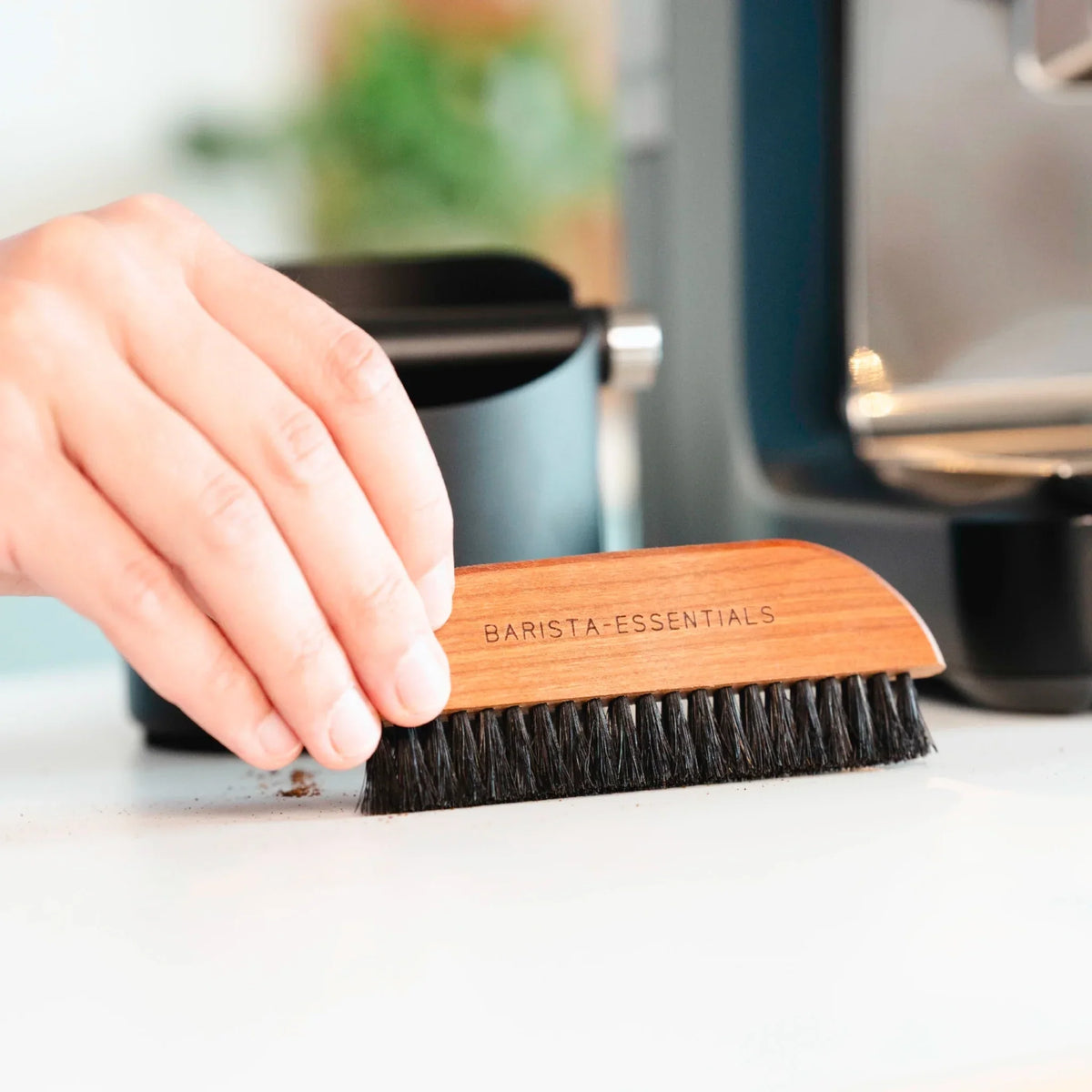 Barista Essentials reinigingsborstel veegt koffieresten naast espressomachine – onderdeel van reinigingsset voor thuis-barista’s