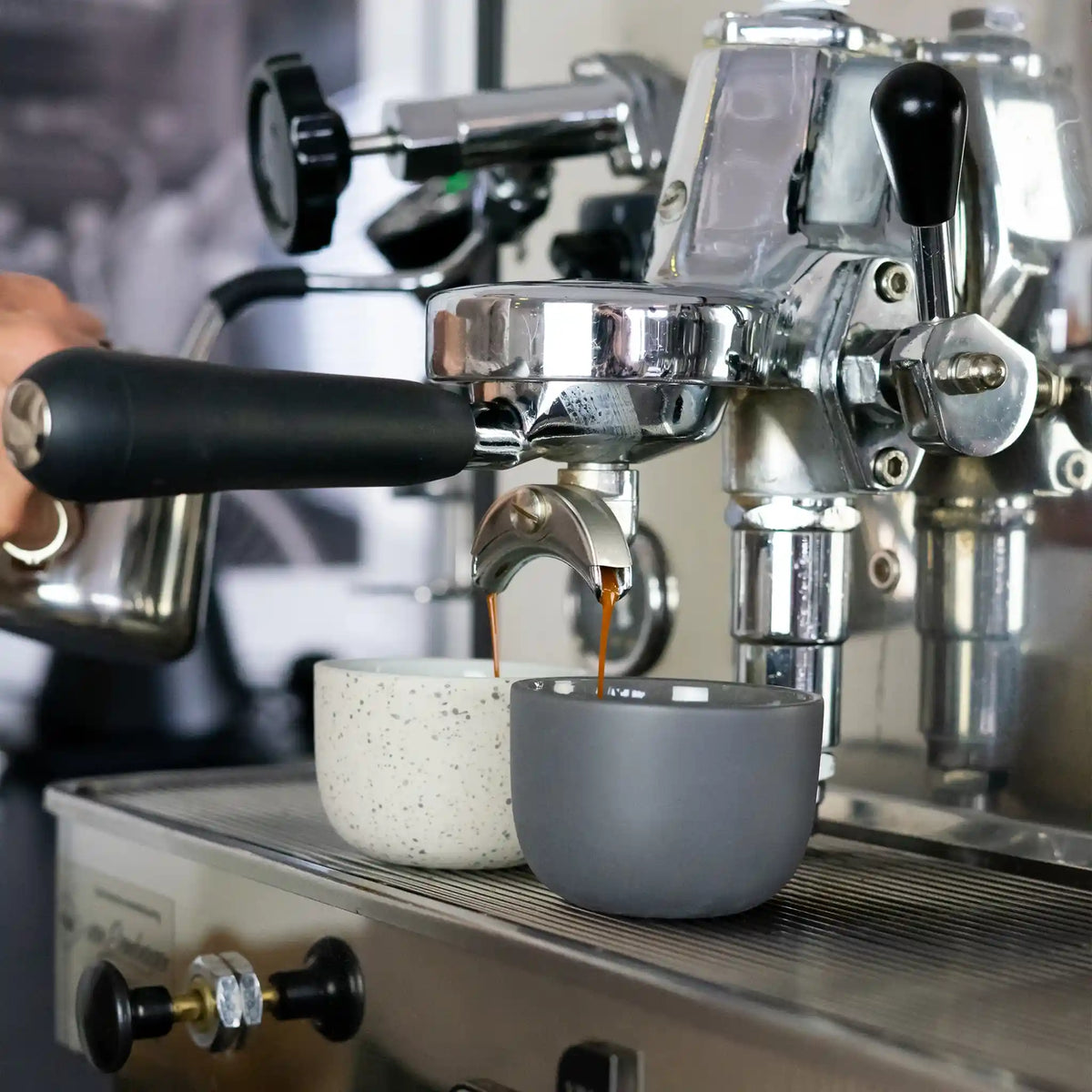 Duurzame barista-tools voor thuis naast een espressomachine, met RVS melkkan, koffieweegschaal, keramische kopjes en vacuüm voorraadpot.