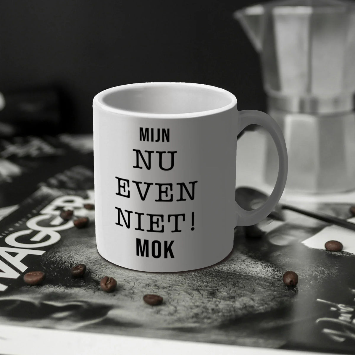 Witte keramische koffiemok met de tekst Mijn nu even niet op een magazine met koffieboontjes en een Bialetti moka pot op de achtergrond