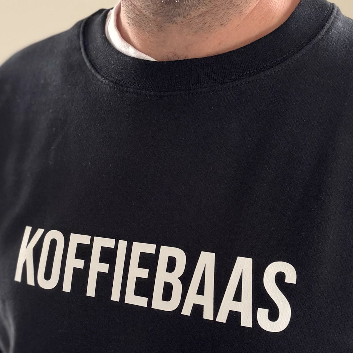 Close-up van witte “KOFFIEBAAS” tekst op zwart sweatshirt – modern barista design