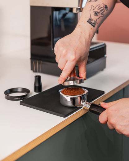 Hand met houten tamper boven een filterdrager met gemalen koffie