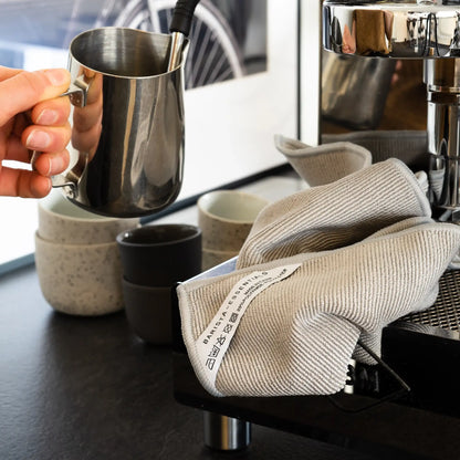 Lichtgrijze doek naast melkkan en kopjes bij een espressomachine