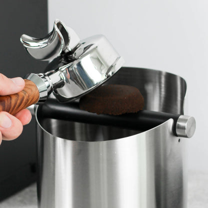 Hand houdt een portafilter boven de beklede stang van een zilver uitklopbak