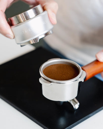 Zilverkleurige tamper en verdeler boven een vlak getampte koffiepuck