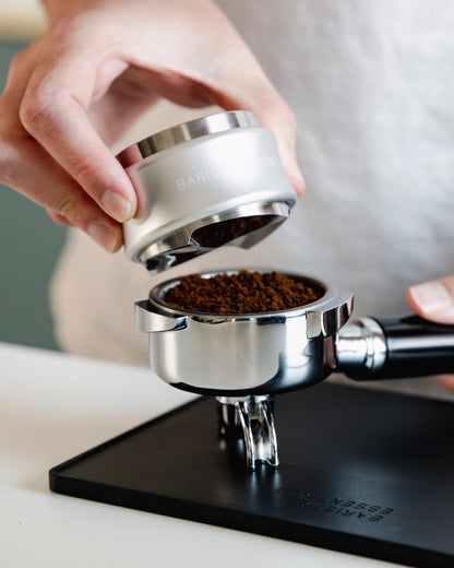 Zilverkleurige tamper en verdeler boven een filterbakje met losse gemalen koffie