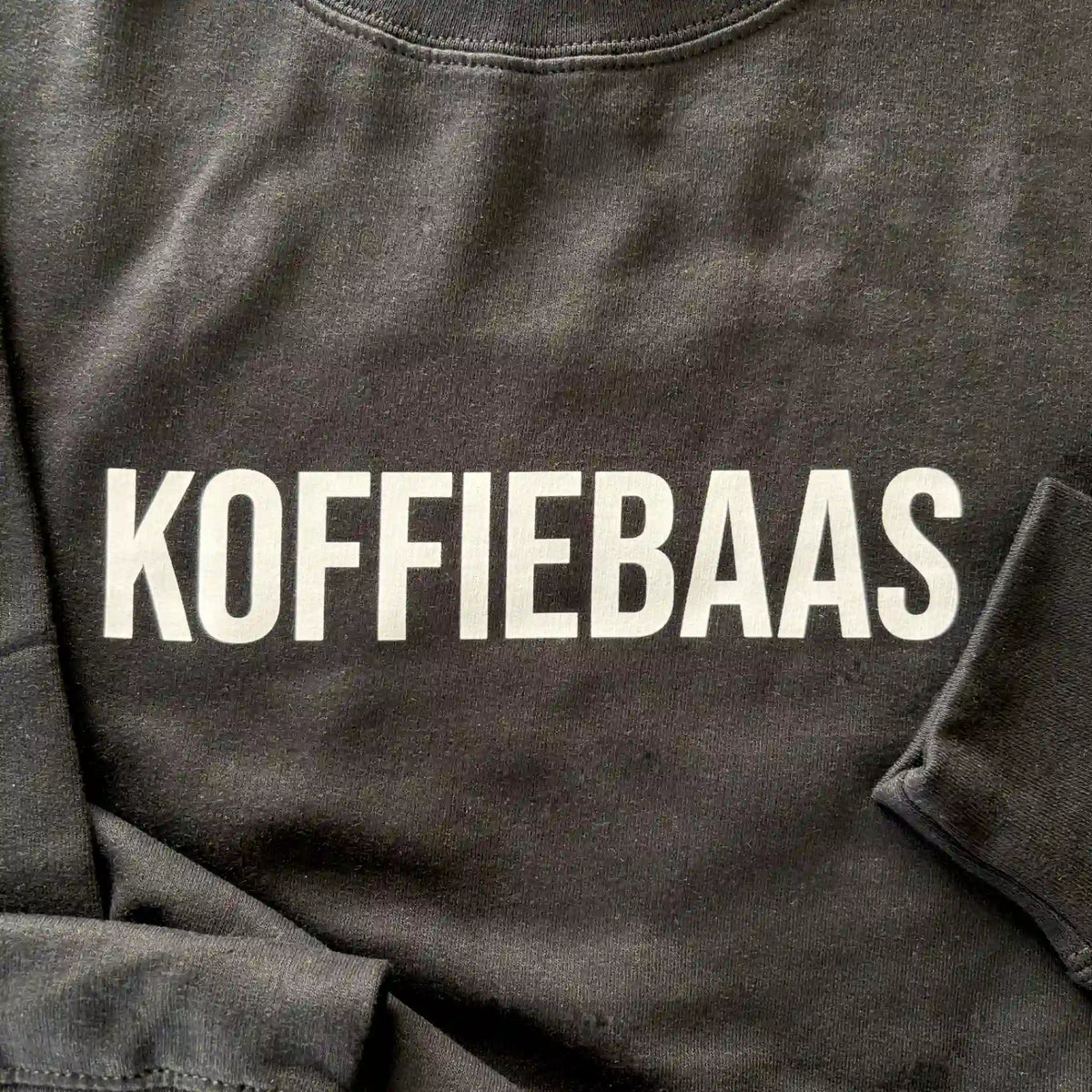 Close-up van de witte “KOFFIEBAAS” print op zwart barista sweatshirt